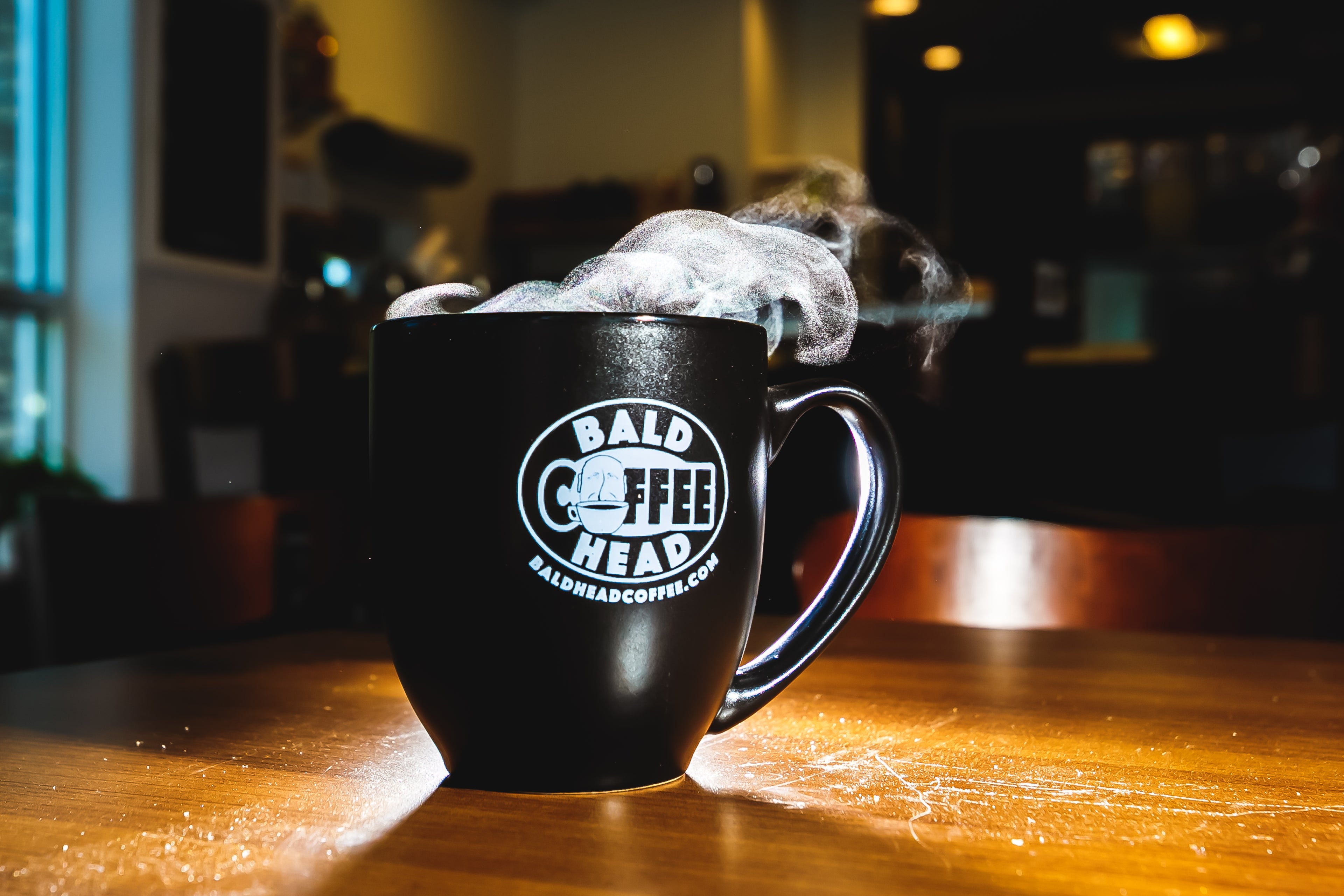 Black mug with 'Bald Coffee Head' logo on a wooden table, steam rising